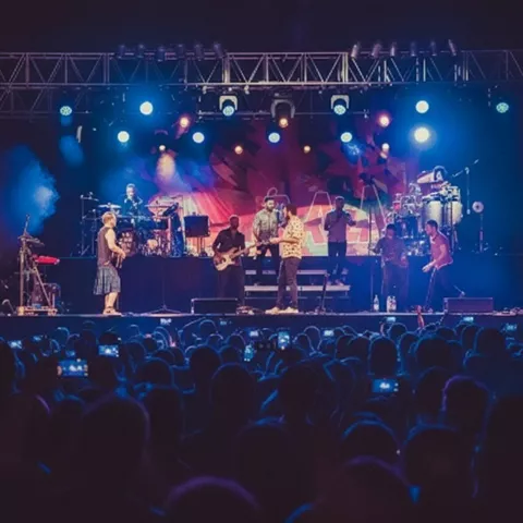 Banda actuando en escenario iluminado ante público con teléfonos móviles.