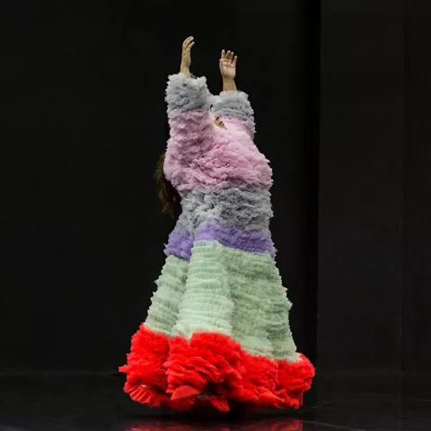 Persona con vestido voluminoso de colores levantando los brazos en un escenario oscuro.