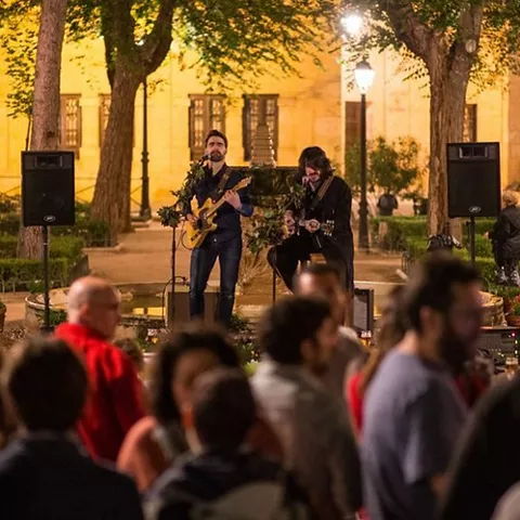 Músicos tocando en una plaza arbolada por la noche ante un público.