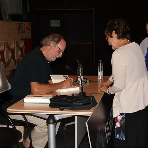 Escritor firmando libros a asistentes en una mesa durante un encuentro cultural.