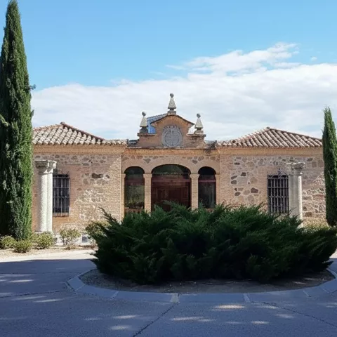 Edificio de piedra con pórtico de arcos y cipreses frente a rotonda ajardinada.