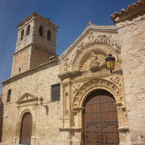 Fachada histórica de piedra con torre y portada ornamentada en una plaza soleada.