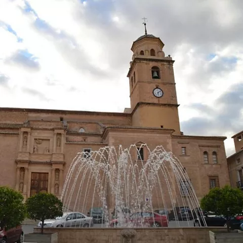 Iglesia de piedra con torre y reloj al fondo, con fuente de agua en primer plano.