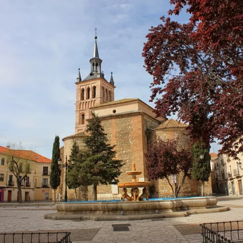 Iglesia de ladrillo y piedra con fuente circular en primer plano y árboles con hojas rojizas.