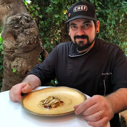 Cocinero con gorra negra presenta un plato en una terraza ajardinada
