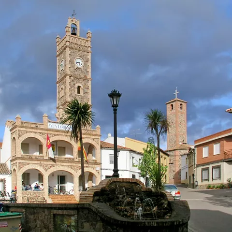 Plaza con ayuntamiento, fuente y torre de iglesia bajo cielo oscuro