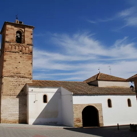 Vista general de una iglesia de muros blancos y tejados de teja, destacando su alta torre de ladrillo contra un cielo azul con nubes ligeras.