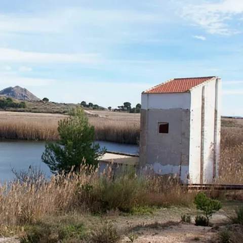 Canal de agua entre juncos con una caseta blanca a la derecha y montes al fondo.