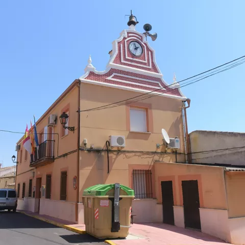 Fachada de edificio municipal con reloj, balcón y banderas en una calle estrecha