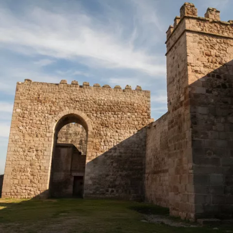 Muralla con arco de acceso y torre de piedra, con sombras alargadas sobre el suelo.