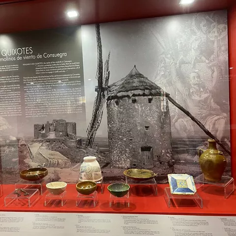 Vitrina de museo con cerámicas antiguas frente a mural en blanco y negro de un molino tradicional.
