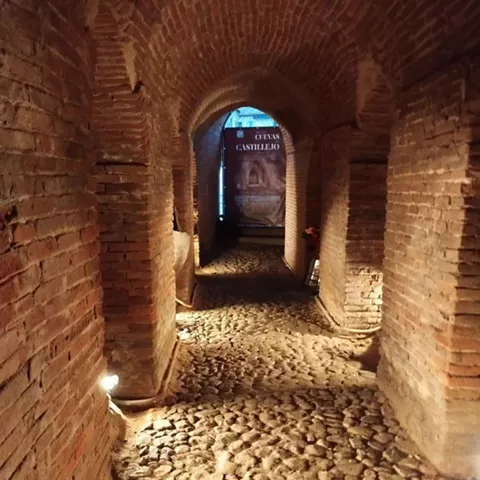 Corredor subterráneo con muros de ladrillo y suelo empedrado iluminado.