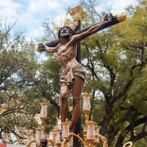 Escultura de Cristo crucificado en paso procesional rodeado de candelabros dorados y flores rojas al aire libre.