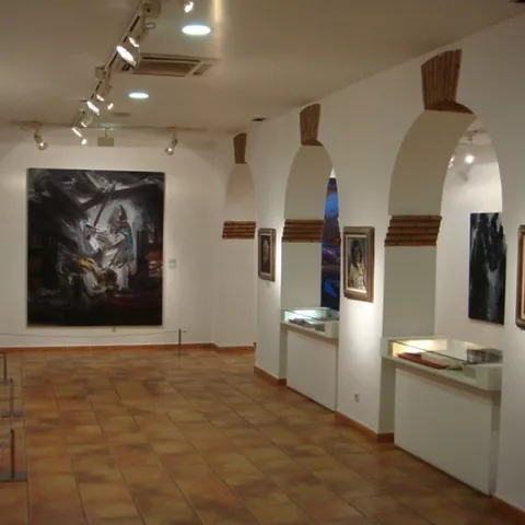 Sala de exposiciones con pinturas en paredes blancas y vitrinas iluminadas.