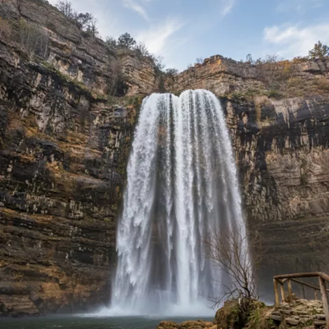 Salto de agua amplio cayendo a una laguna oscura, con barandilla junto a la orilla.