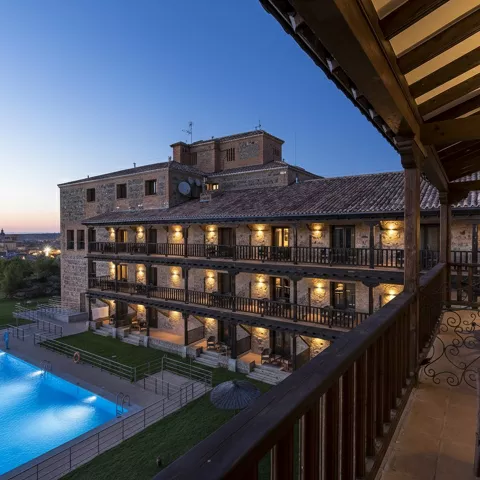 Fachada iluminada de hotel con galerías y piscina al anochecer.