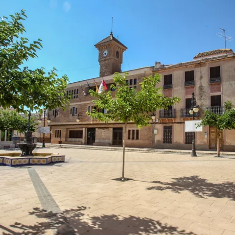 Plaza amplia con edificio institucional, fuente central y árboles.
