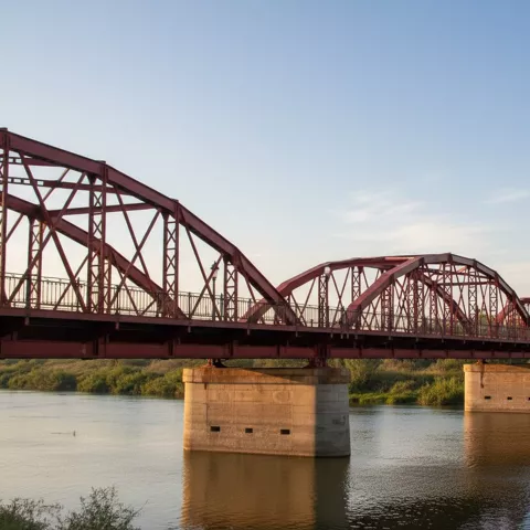 Vista lateral de un puente de hierro rojo sobre el agua, con vegetación en la orilla.