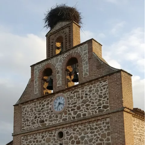 Una vista de cerca de la torre del reloj de una iglesia de piedra y ladrillo con campanas visibles y un nido de cigüeña en la parte superior.
