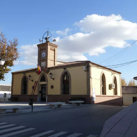 Edificio municipal de color amarillo pálido con grandes ventanas góticas y una torre de reloj central de ladrillo coronada por una estructura de metal para campanas y altavoces, situado frente a una plaza adoquinada.