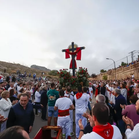 Plaza abarrotada con un paso con cruz y flores, con asistentes aplaudiendo alrededor.