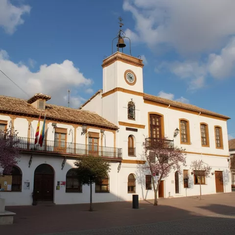 Un edificio municipal blanco de dos plantas con un campanario y un reloj, frente a una plaza adoquinada con árboles.