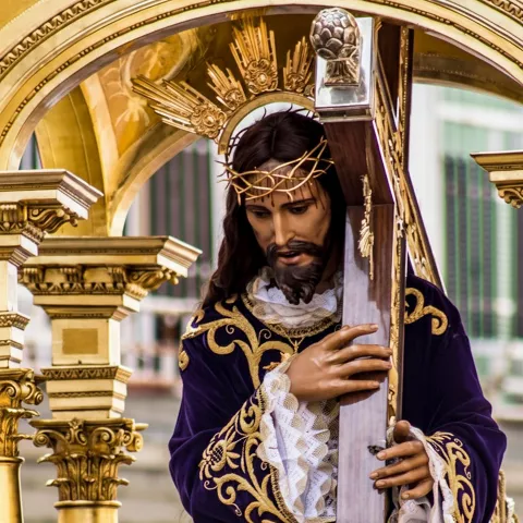 Imagen religiosa con túnica morada y cruz de madera bajo arco dorado.