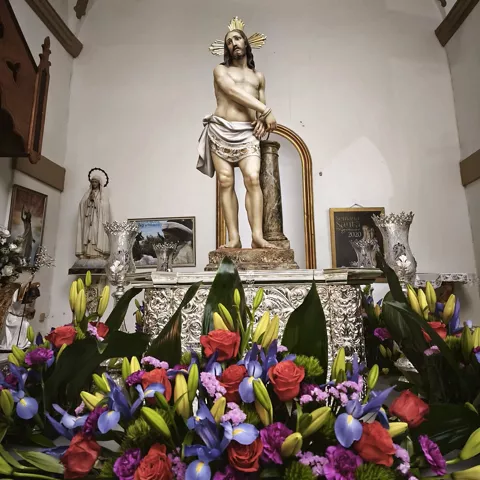 Imagen religiosa en el interior de un templo, rodeada de flores y elementos ornamentales.
