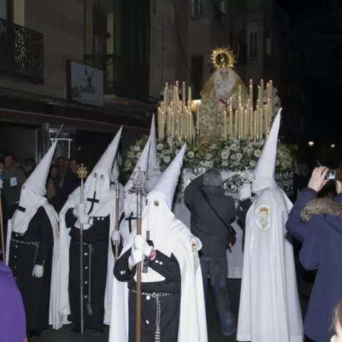 Procesión nocturna con penitentes encapuchados junto a un paso con velas y flores.