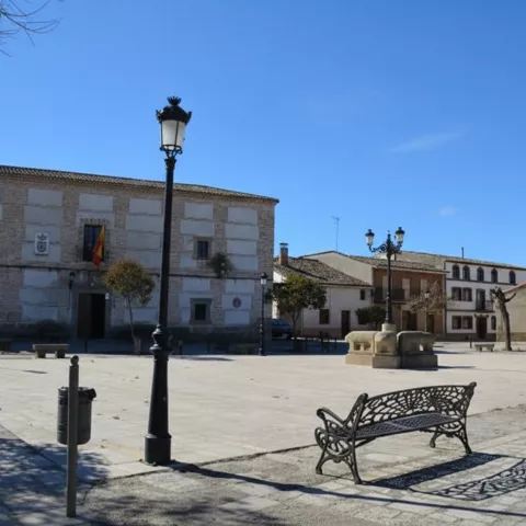 Vista amplia de una plaza de pueblo pavimentada que incluye un edificio administrativo con banderas, bancos de hierro forjado, farolas clásicas y una iglesia de piedra a la derecha.