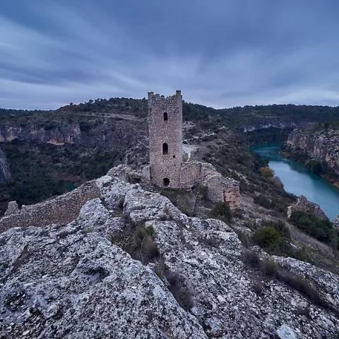 Torre de piedra en lo alto de un risco junto a un río en paisaje rocoso.