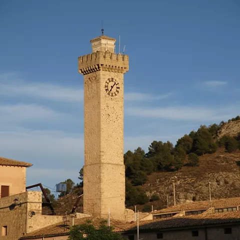 Torre cuadrada con reloj y colinas arboladas al fondo.
