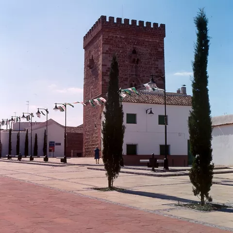 Plaza con torre almenada y edificio blanco, banderines y cipreses.