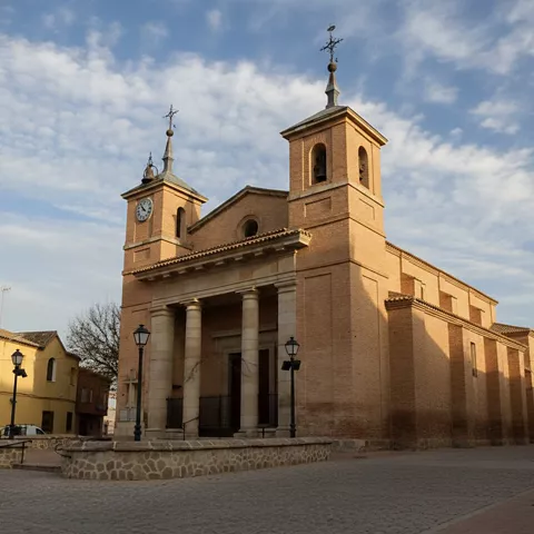 Una iglesia de ladrillo con un pórtico de columnas de piedra y dos torres campanario simétricas bajo un cielo parcialmente nublado