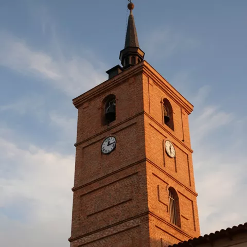 Primer plano de una torre campanario de ladrillo rojo iluminada por la luz del atardecer, destacando sus campanas y relojes.