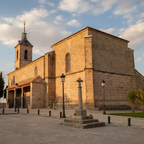 Una iglesia de piedra rústica con un campanario y una cruz de piedra en el patio delantero, con árboles y farolas que la enmarcan.