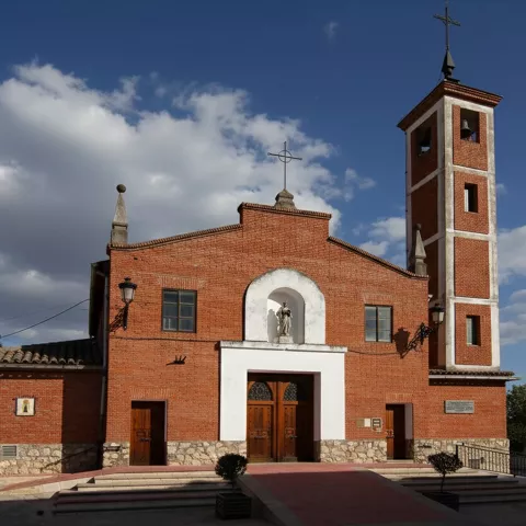 Fachada frontal de una iglesia de ladrillo rojo con una torre campanario alta y estrecha a la derecha y una estatua en un nicho sobre la puerta principal.