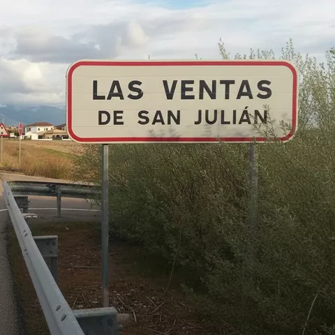 Señal de tráfico rectangular blanca con borde rojo que indica la entrada a una localidad, situada al borde de una carretera recta con árboles y montañas al fondo.