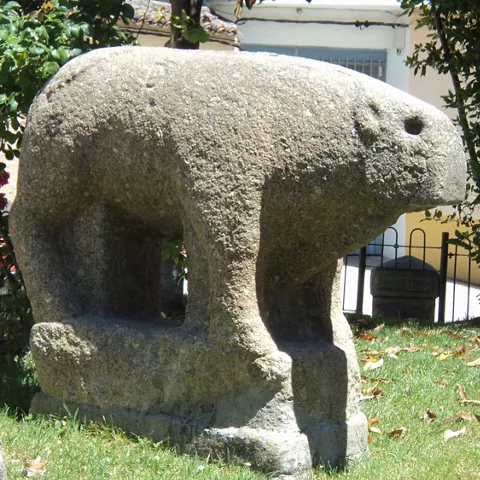 Escultura zoomorfa de piedra sobre pedestal en una zona ajardinada.