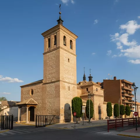 Vista de una iglesia de ladrillo y piedra con una torre campanario alta, ubicada en una calle de un pueblo bajo un cielo parcialmente nublado.