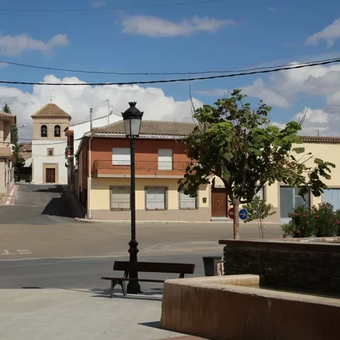 Gemini ha dicho Aquí tienes los textos alternativos breves y descriptivos para las imágenes proporcionadas:  image_0.png: Una toma a nivel de calle de una plaza de un pequeño pueblo español, con un banco de madera, una farola y una fuente de piedra en primer plano