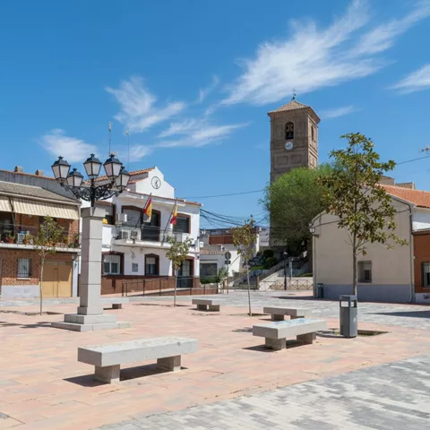 Una plaza de pueblo adoquinada con una fuente ornamental de varios niveles y un campanario de iglesia que se asoma detrás de los árboles.