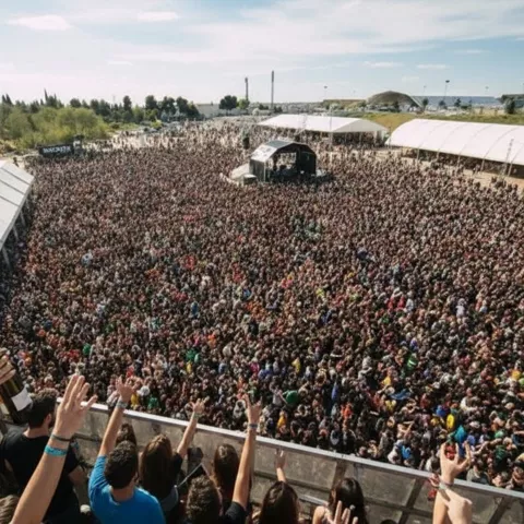 Vista diurna desde una plataforma con miles de asistentes concentrados frente a un escenario al fondo.