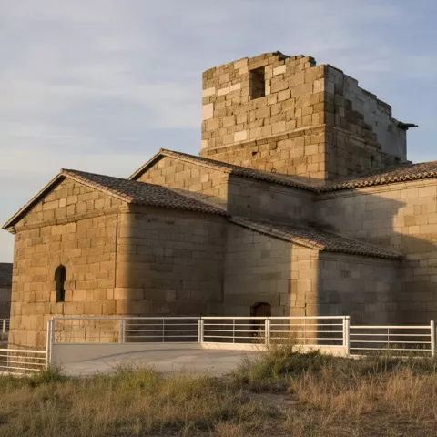 Lateral de la iglesia de piedra con torre cuadrada, junto a hierba seca y barandilla blanca.