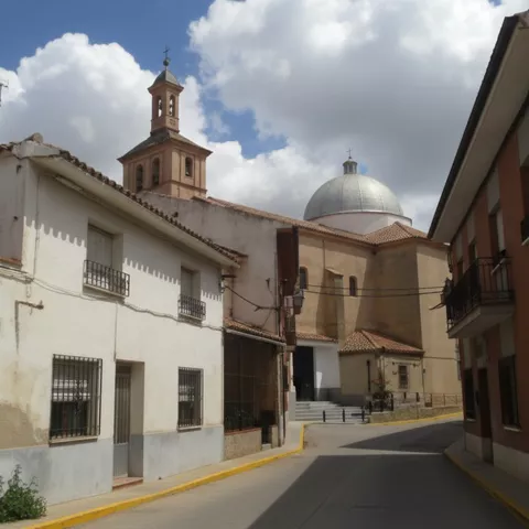 Una calle curva de un pueblo con bordillos amarillos, donde casas blancas conducen a una iglesia con una cúpula plateada y una torre de ladrillo bajo un cielo azul con nubes.