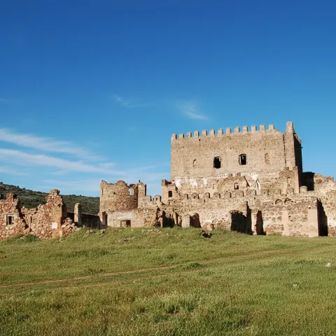 Un plano general muestra las ruinas de piedra de una gran fortaleza medieval situada sobre una colina cubierta de hierba verde, bajo un cielo azul despejado.