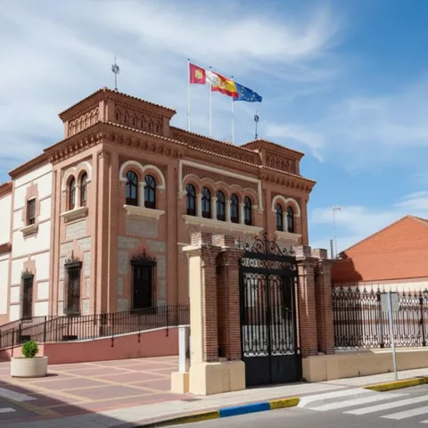 Vista exterior de un edificio de ladrillo rojo y piedra, con balcones de hierro forjado, tres banderas en el tejado y un patio delantero.