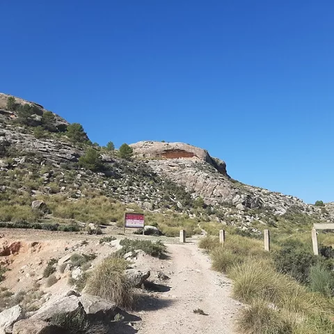 Camino de tierra hacia un abrigo rocoso en la ladera, bajo cielo azul.
