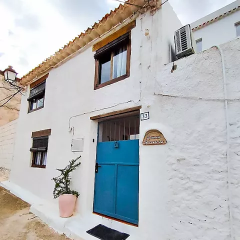 Fachada de casa blanca con puerta azul y ventanas pequeñas en calle estrecha.