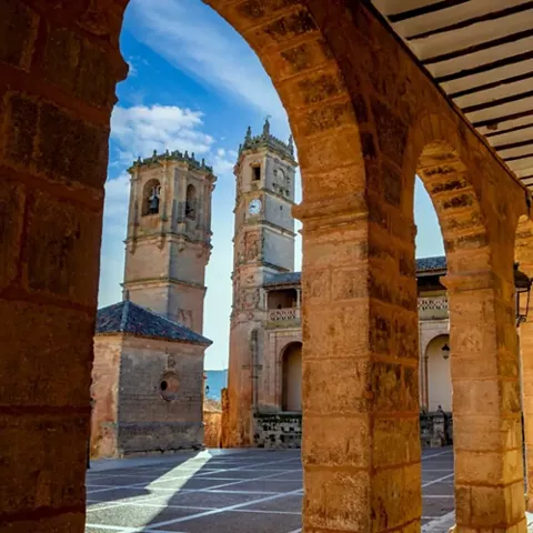 Galería porticada de piedra con torres al fondo y luz lateral.